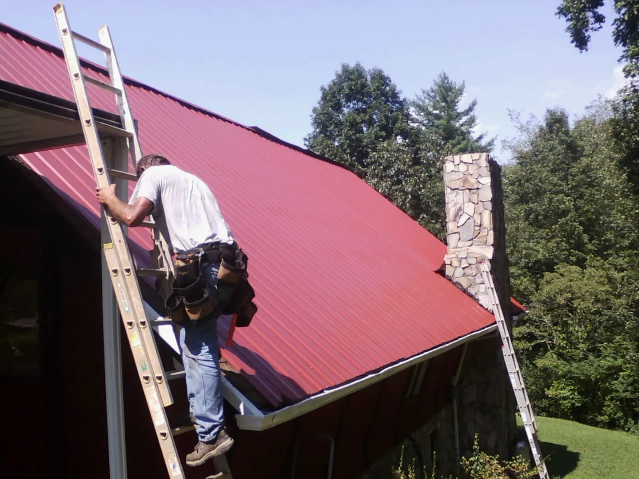 Roofer on a ladder beside a red metal roof and stone chimney — Soffit & Fascia Repair by Roofing Brothers in Tiltonsville